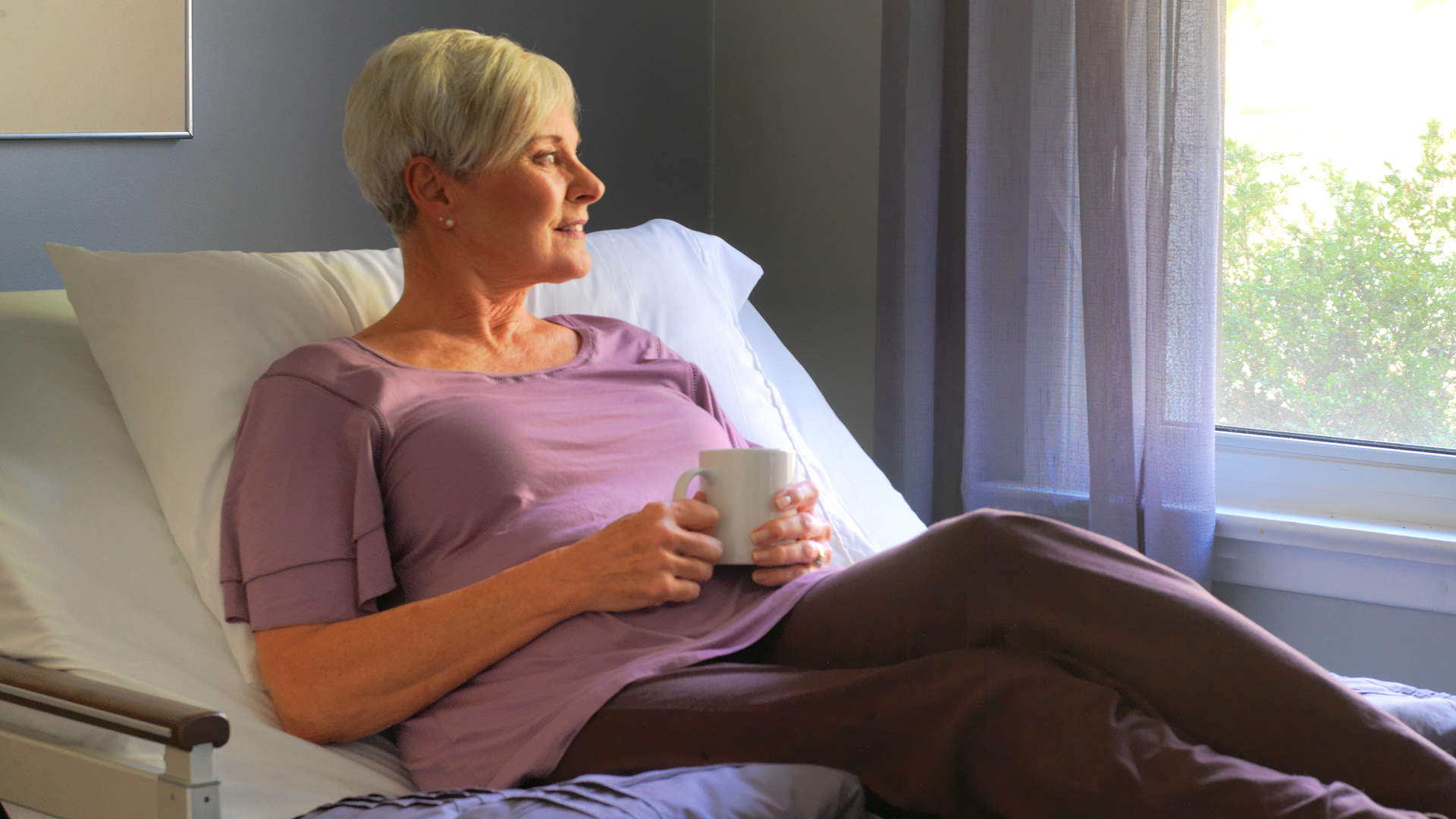 Mature woman with short gray hair smiling, sitting in a hospital bed by a window, holding a white cup.