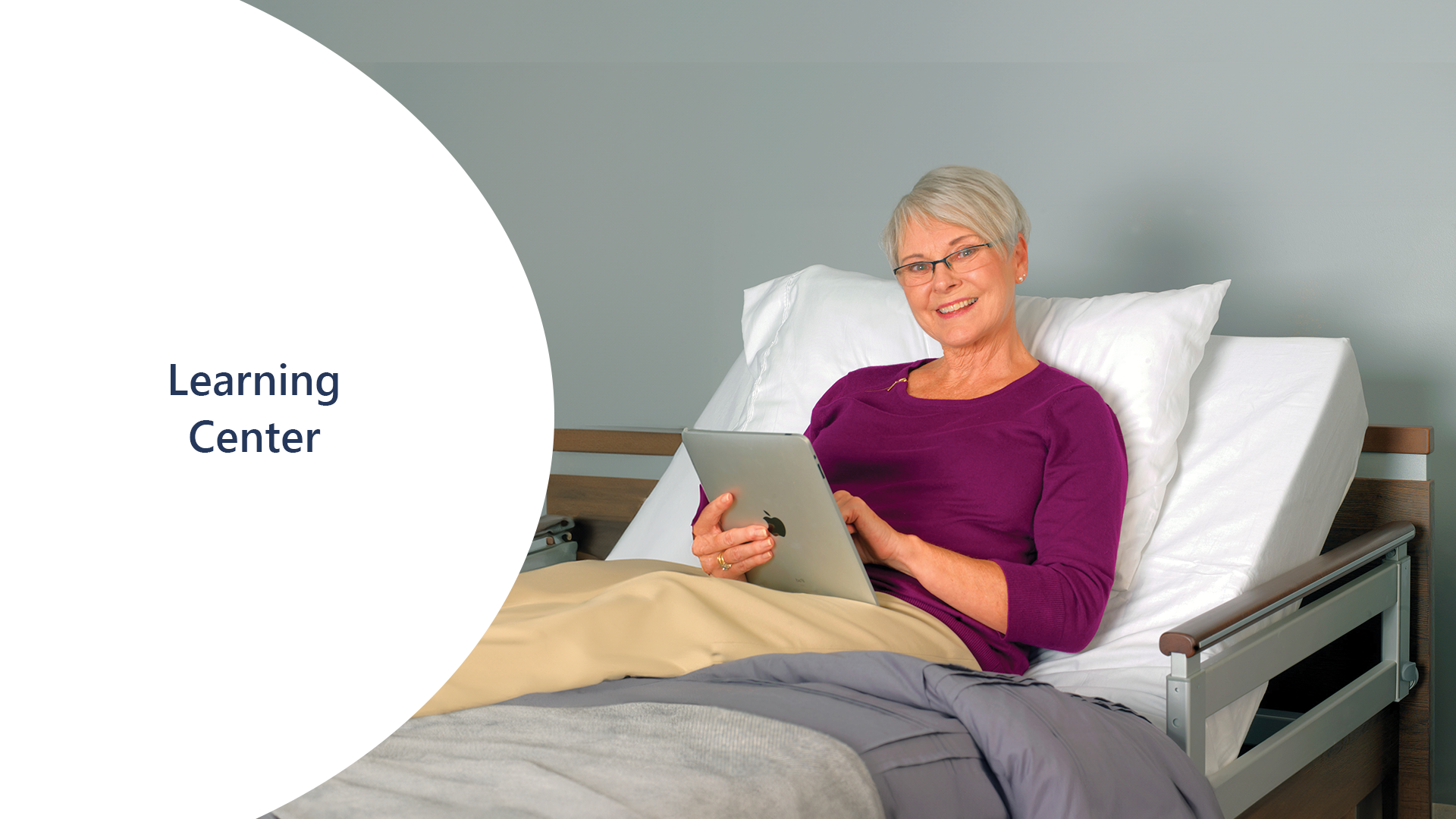 Older woman with short gray hair, wearing glasses and a purple top, sits up in a hospital bed using a tablet. Text on the left reads "Learning Center".