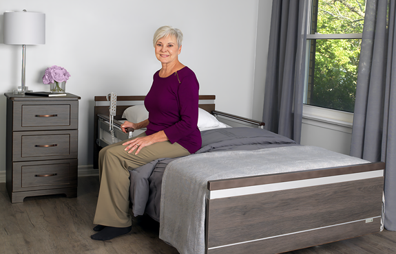 An older woman sits on the edge of a bed in a neatly arranged bedroom with gray furnishings, holding a bed assist rail.