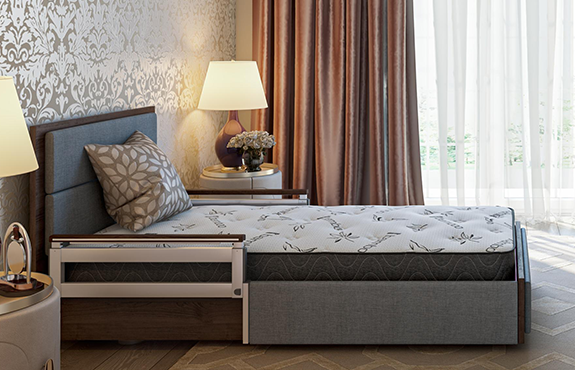 A neatly made single bed with safety rails in a well-lit bedroom, beside a nightstand with a lamp and flowers, and large curtained windows.