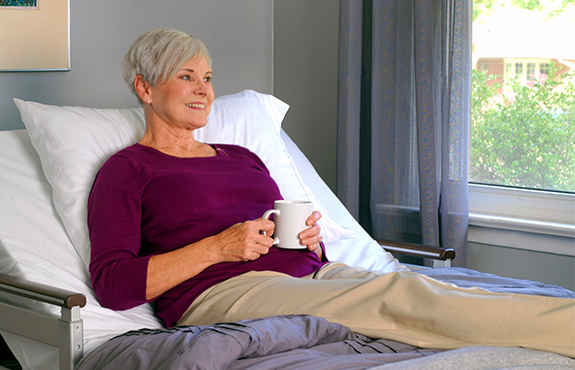 Older woman with short gray hair, wearing a purple top and beige pants, sits up in bed holding a white mug, near a window with light coming in.